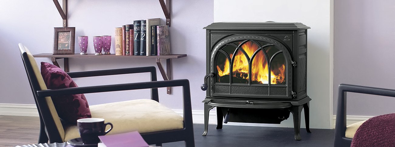 Czarny, żeliwny piec wolnostojący Jotul F 400 z paleniskiem, ustawiony w salonie z drewnianymi meblami i dekoracjami. W tle widać książki na półkach i wygodny fotel.